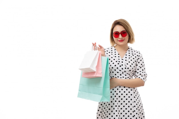 Foto gratuita una giovane bella donna di vista frontale in vestito a pois in bianco e nero in occhiali da sole rossi che tengono posa dei pacchetti di acquisto