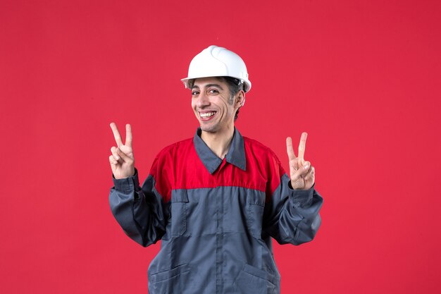 Front view of young builder in uniform wearing hard hat making victory gesture on isolated red wall