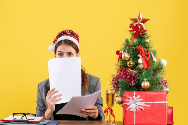 Вид спереди молодая бизнес-леди в рождественской шляпе сидит за столом, закрывая лицо документами, рождественской елкой и подарочным коктейлем