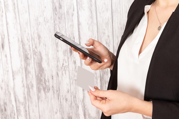 Front view young businesswoman in strict clothes black jacket holding card and phone on light-white surface