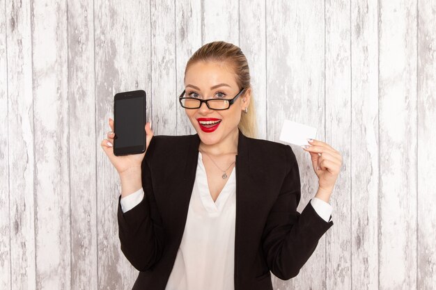 Front view young businesswoman in strict clothes black jacket holding card and phone on the white surface