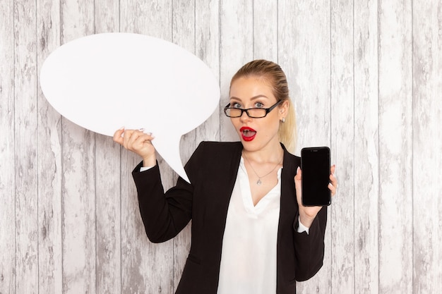 Front view young businesswoman in strict clothes black jacket holding her phone and white sign on the white surface