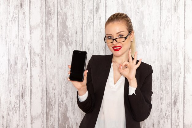 Front view young businesswoman in strict clothes black jacket holding her phone with smile on white surface