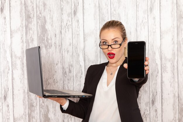 Front view young businesswoman in strict clothes black jacket using her laptop and holding phone on white surface