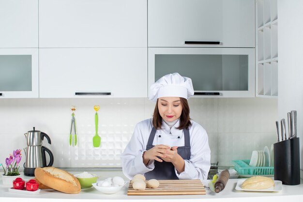 Front view of young busy female chef in uniform standing behind table preparing pastry in the white kitchen