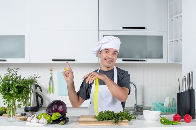 Front view young chef in uniform pointing at ceiling in modern kitchen