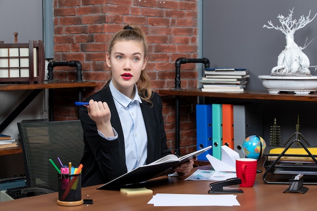 彼女の机に座って、オフィスでドキュメントを保持している若い集中女性アシスタントの正面図