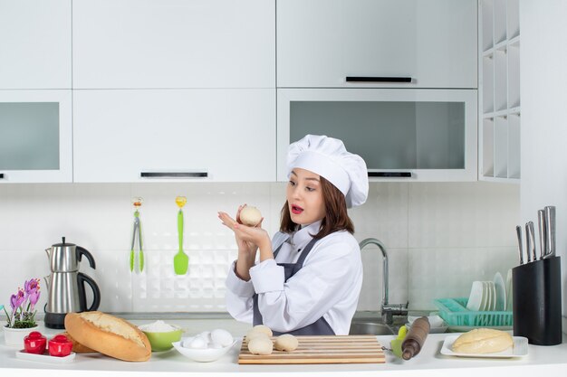 Front view of young concentrated female chef in uniform standing behind table preparing pastry in the white kitchen