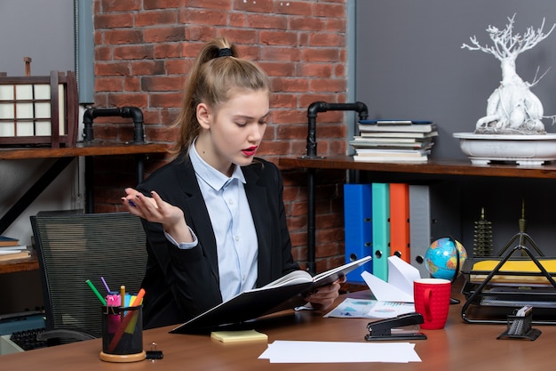Вид спереди молодой уверенной женщины-помощника, сидящей за своим столом и проверяющей документ в офисе