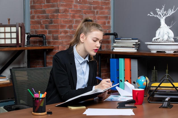 彼女の机に座って、オフィスでドキュメントを保持している若い自信を持って女性アシスタントの正面図