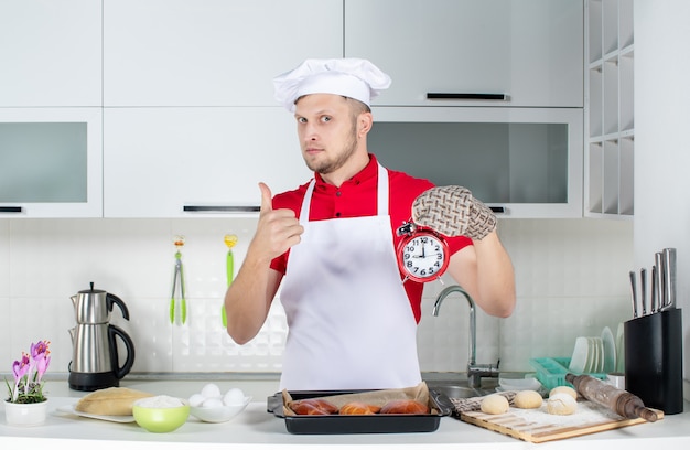 Вид спереди молодого уверенного в себе шеф-повара в держателе, держащего часы и делающего жест на белой кухне