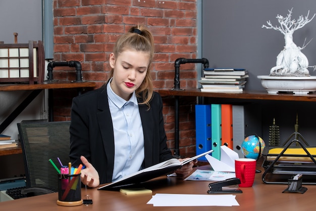 Front view of young confused female assistant sitting at her desk and holding document in the office