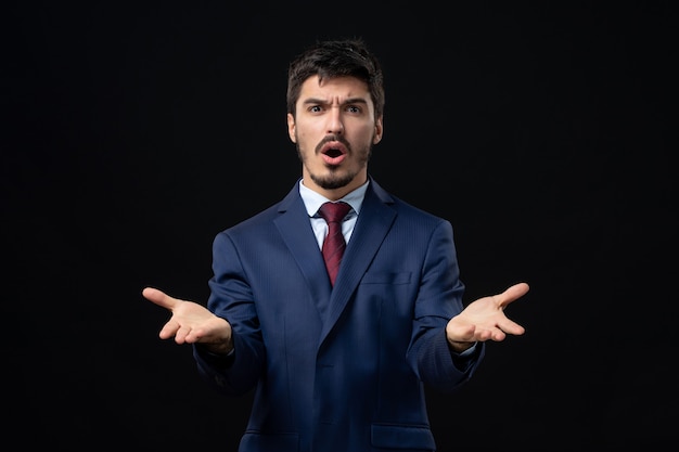 Front view of young confused man in suit asking something with suspicious facial expression on isolated dark wall