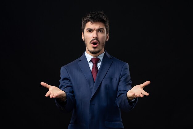 Front view of young confused man in suit asking something with suspicious facial expression on isolated dark wall