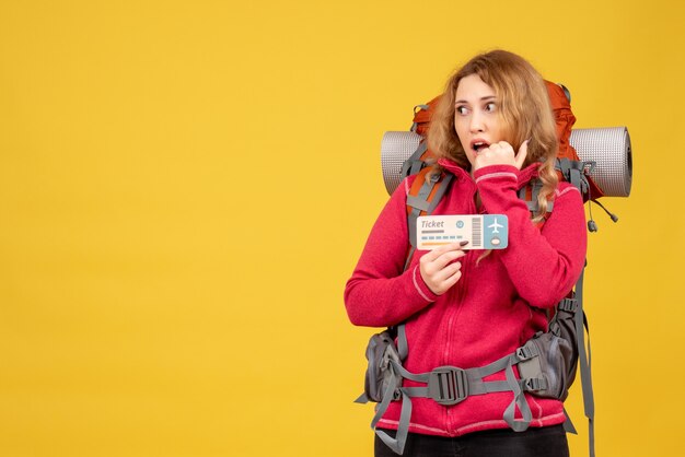 Front view of young confused travelling girl in medical mask holding ticket and pointing up