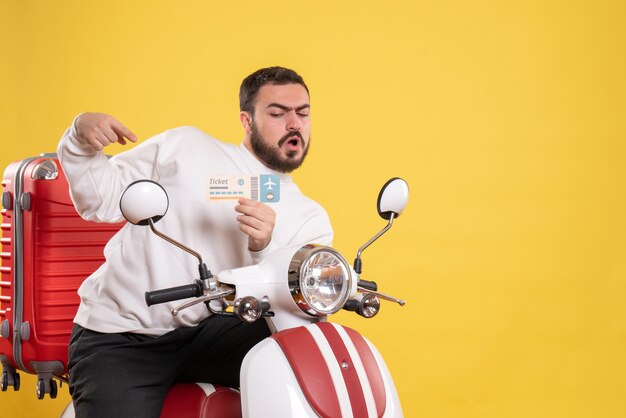 Front view of young confused travelling man sitting on motorcycle with suitcase on it holding ticket on isolated yellow background