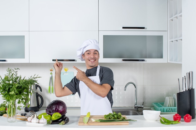 Front view young cook in uniform pointing at cupboard in kitchen