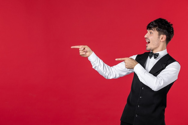 Front view of of young curious male waiter in a uniform with bowtie on neck and pointing up on the right side with both hands on red wall