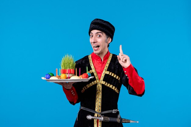 Front view of young curious man in traditional dress holding tray filled with national confectionery pointing up on blue background