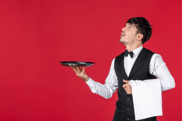 Front view of of young curious man waiter in a uniform with bowtie on neck holding tray and towel looking up on red wall