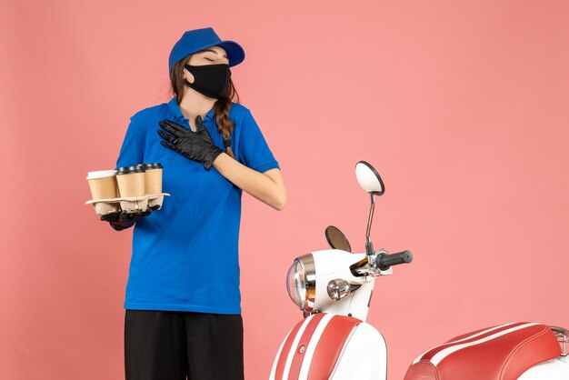 Front view of young dreamy courier girl wearing medical mask gloves standing next to motorcycle holding coffee small cakes on pastel peach color background