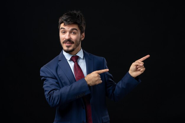 Front view of young emotional smiling man in suit pointing up on isolated dark wall