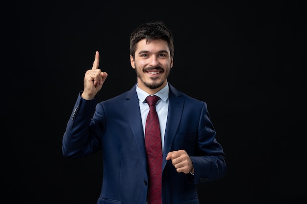 Front view of young emotional smiling man in suit pointing up on isolated dark wall