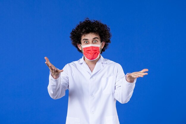 正面図青い背景に赤いマスクで医療スーツを着た若い興奮した医者薬ワクチン健康コビデミック医学病院ウイルス