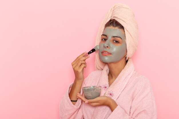 Front view young female after shower in pink bathrobe applying mask on pink surface