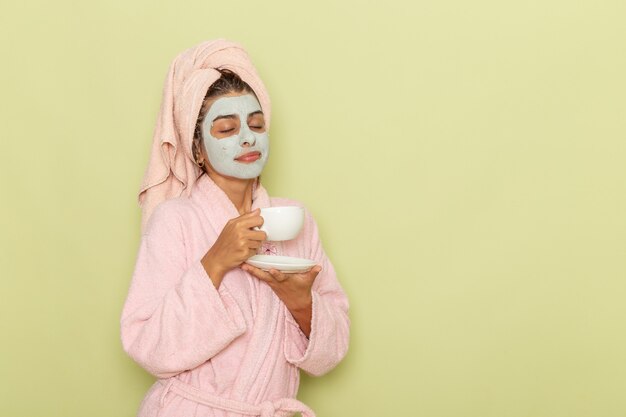 Front view young female after shower in pink bathrobe drinking coffee on green surface