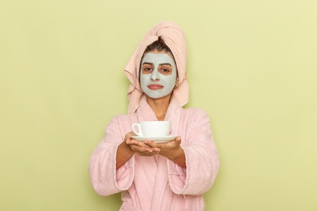 Вид спереди молодая женщина после душа в розовом халате пьет кофе на светло-зеленой поверхности