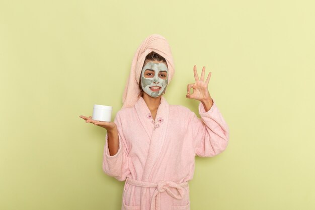 Front view young female after shower in pink bathrobe holding cream and smiling on a green surface