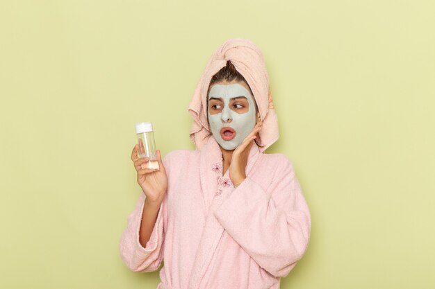 Front view young female after shower in pink bathrobe holding make-up remover on green surface