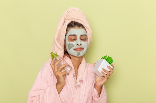 Вид спереди молодая женщина после душа в розовом халате, держащая средства для снятия макияжа на зеленом полу, крем-маска, душ, уход за собой, косметическая ванна