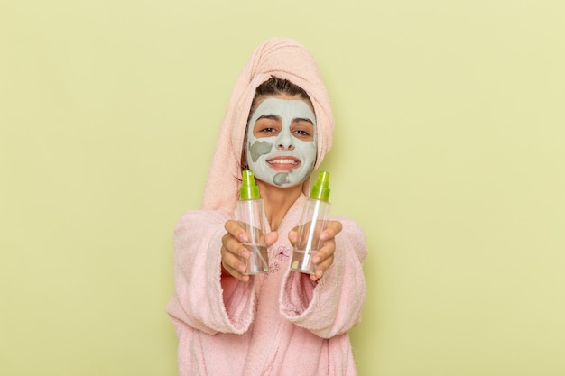 Вид спереди молодой женщины после душа в розовом халате, держащей средства для снятия макияжа на зеленой поверхности