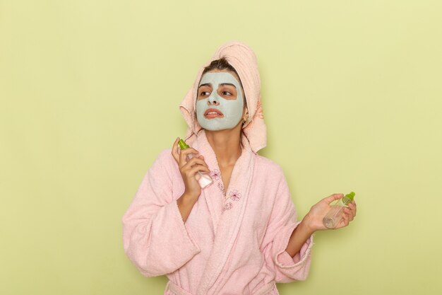 Front view young female after shower in pink bathrobe holding sprays on green desk