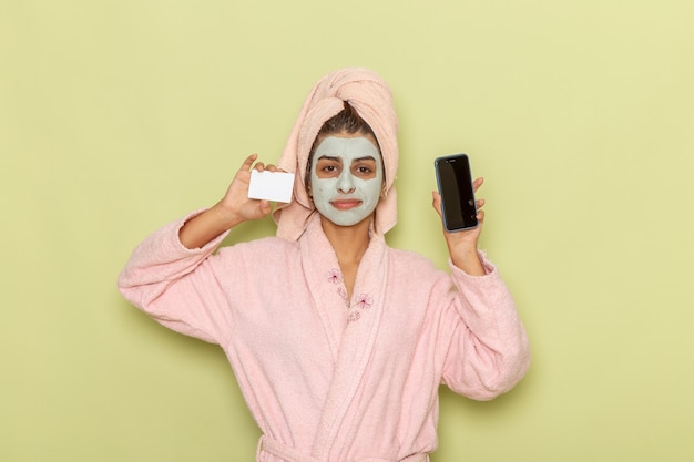 Front view young female after shower in pink bathrobe holding white card and phone on green desk