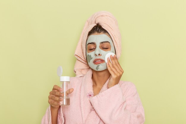 Front view young female after shower in pink bathrobe using make-up remover on a green surface