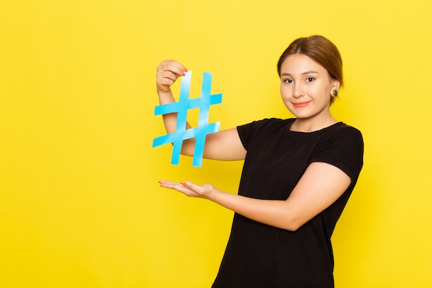 Free photo a front view young female in black dress holding blue sign smiling on yellow