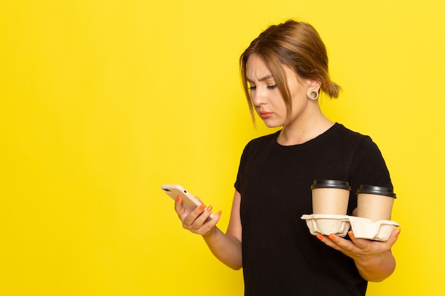 A front view young female in black dress holding coffee cups and using a phone on yellow