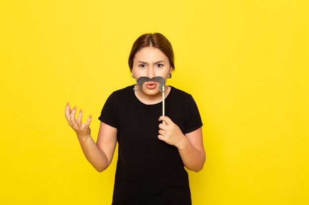 A front view young female in black dress posing with fake mustache with displeased expression on yellow