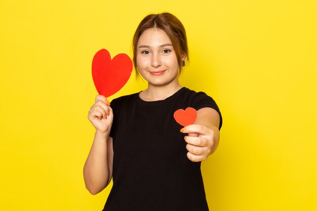 A front view young female in black dress posing with red heart shapes smiling on yellow