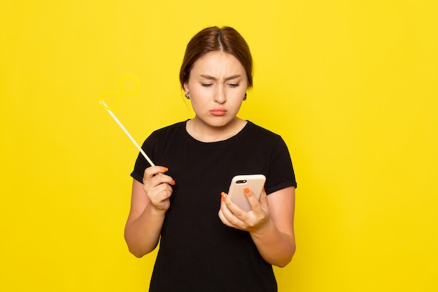 A front view young female in black dress posing with smartphone and toy stick sunglasses on yellow