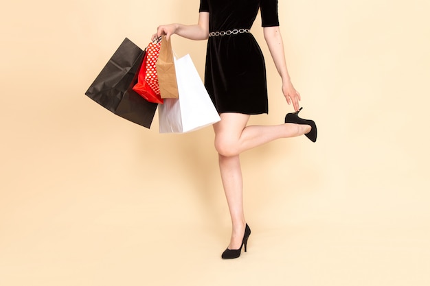 Вид спереди молодая женщина в черном платье с цепным поясом, держащая пакеты с покупками на бежевом