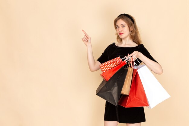 A front view young female in black dress with chain belt holding shopping packages on beige