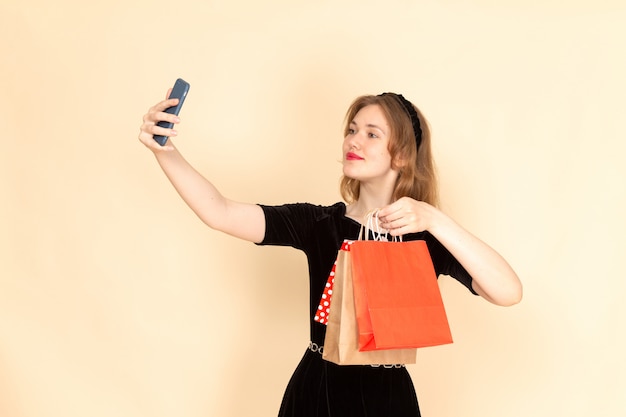 A front view young female in black dress with chain belt holding shopping packages and taking a selfie on beige