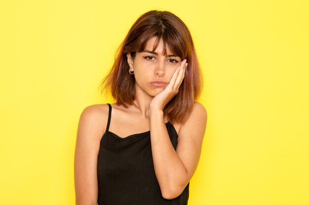 Front view of young female in black shirt and grey jeans having toothache on yellow wall