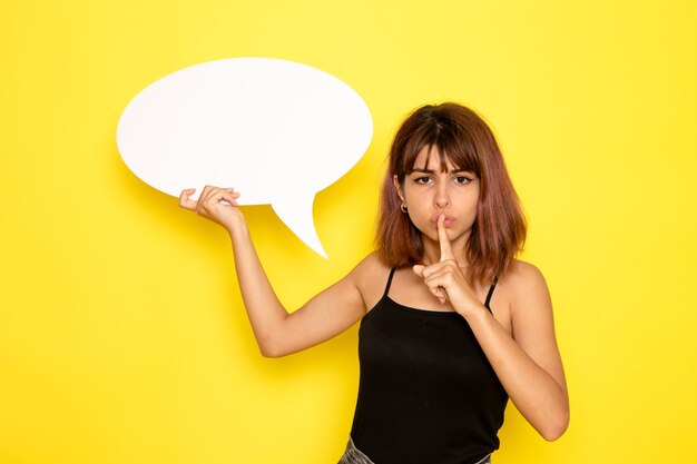 Front view of young female in black shirt and grey jeans holding big white sign on yellow wall