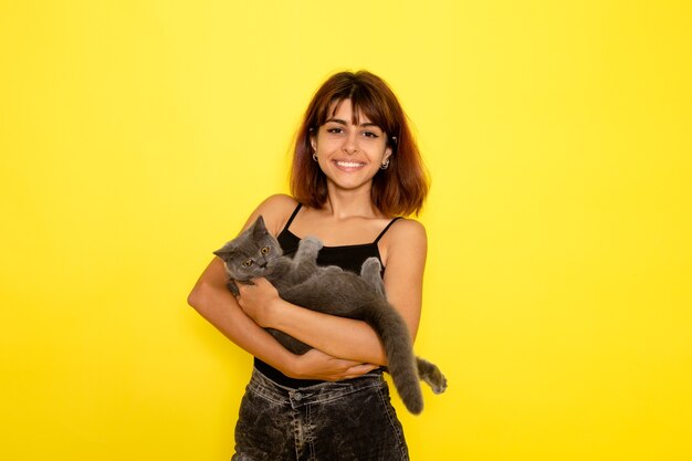 front view of young female in black shirt and grey jeans holding cute grey kitten on yellow wall