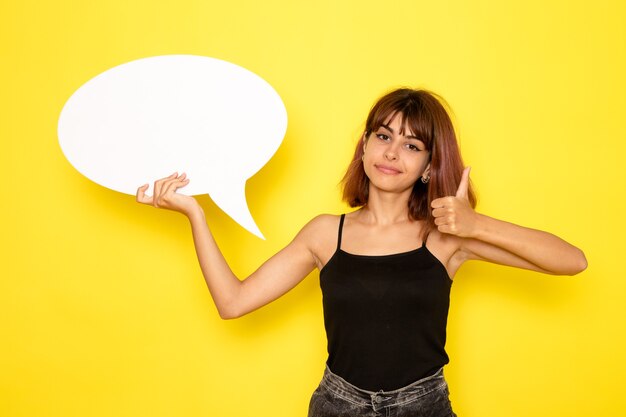 Front view of young female in black shirt and grey jeans holding a huge white sign on the yellow wall